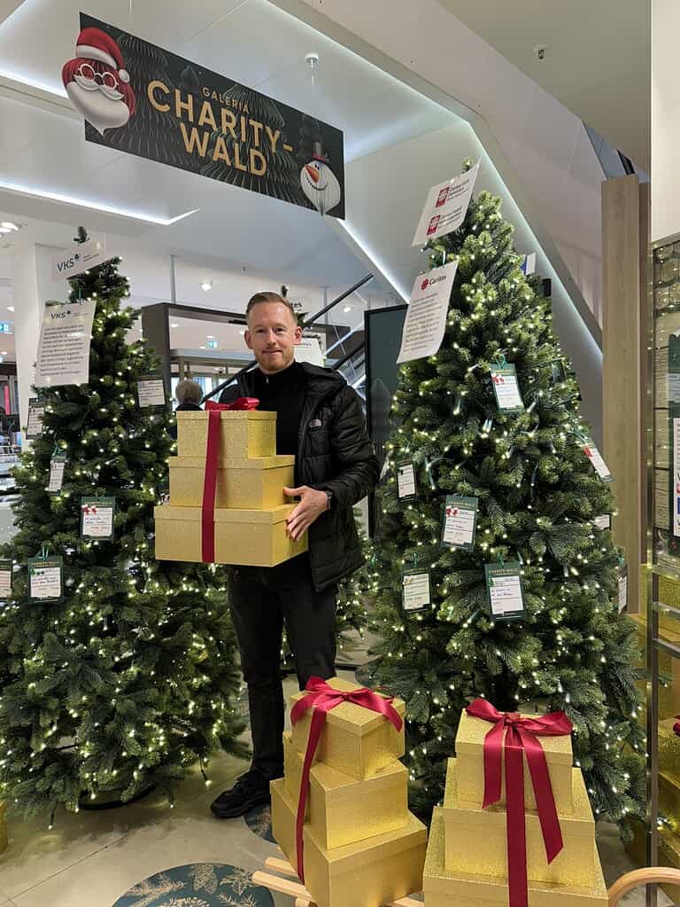 Felix Schönebeck hält mehrere goldene Geschenkboxen mit roten Schleifen vor zwei weihnachtlich geschmückten Tannenbäumen im "Charity-Wald" der Galeria. Die Bäume sind mit Wunschzetteln für eine Spendenaktion behangen.