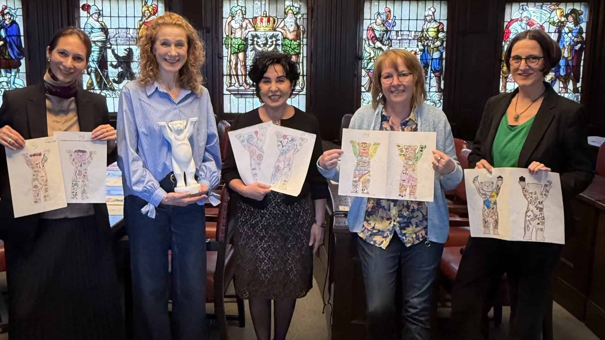 Vier Frauen stehen vor den bunten Fenstern im BVV-Saal im Rathaus Reinickendorf. Drei halten die gezeichneten Entwürfe für die Gestaltung der