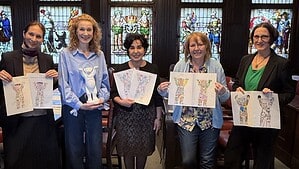 Vier Frauen stehen vor den bunten Fenstern im BVV-Saal im Rathaus Reinickendorf. Drei halten die gezeichneten Entwürfe für die Gestaltung der "Ausbildungsbuddys" in den Händen. Eine Frau hält einen weißen Buddybären.