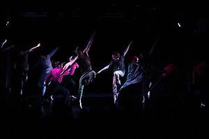 Junge Tänzerinnen und Tänzer der Young Trybe Dance Company in bunten Kostümen bei einer Aufführung auf dunkler Bühne.