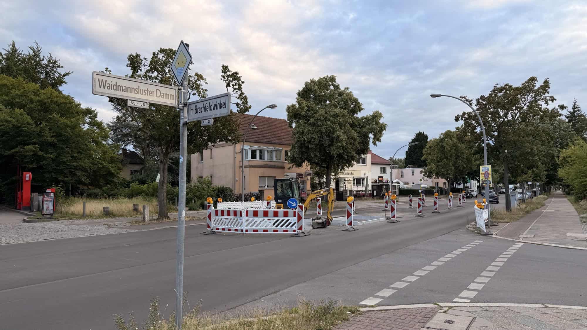 Straßenschild Waidmannsluster Damm Ecke Im Brachfeldwinkel, dahinter Absperrbaken und ein Bagger auf der Fahrbahn