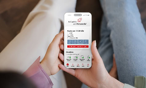 Zwei Hände halten ein Smartphone, auf dem Bildschirm ist das Dashboard der DRK-Blutspende-App mit dem nächsten Spendetermin zu sehen.