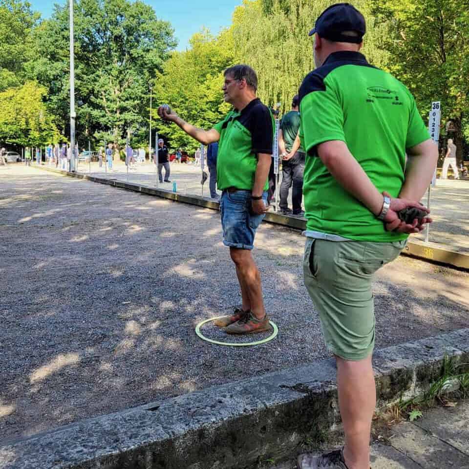 Zwei Spieler in grünen Trikots des Club Bouliste de Berlin auf der Freiluft-Boulebahn, einer wirft eine Kugel aus dem Wurfkreis.