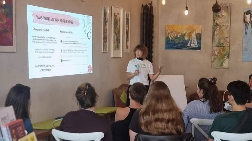 Eine Frau mit Mikrofon spricht vor sitzenden Gästen, neben ihr eine Präsentation an der Wand.