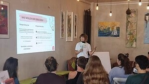 Eine Frau mit Mikrofon spricht vor sitzenden Gästen, neben ihr eine Präsentation an der Wand.