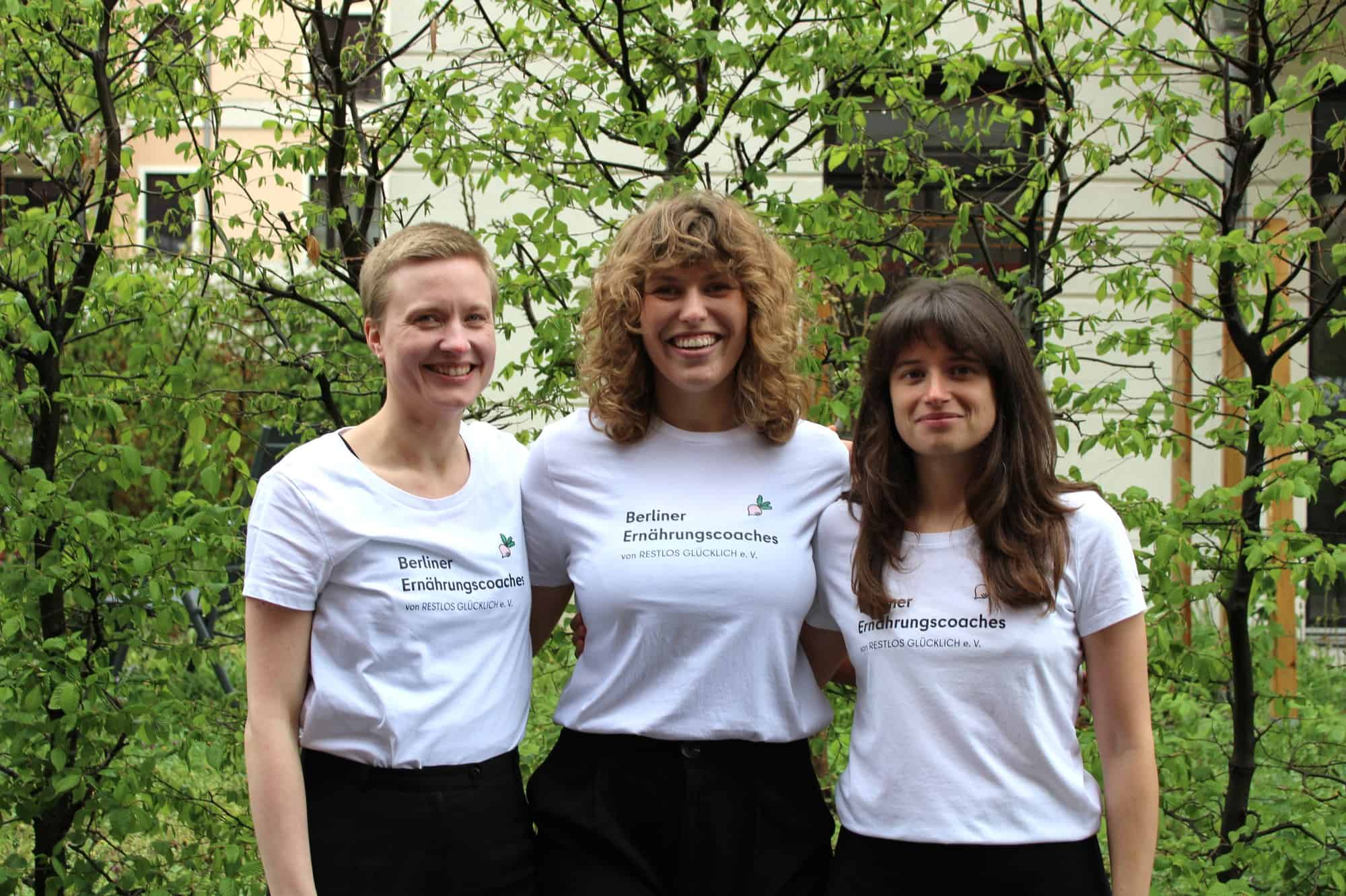 Drei Frauen in weißen T-Shirts mit dem Aufdruck Berliner Ernährungscoaches stehen vor grünen Bäumen.