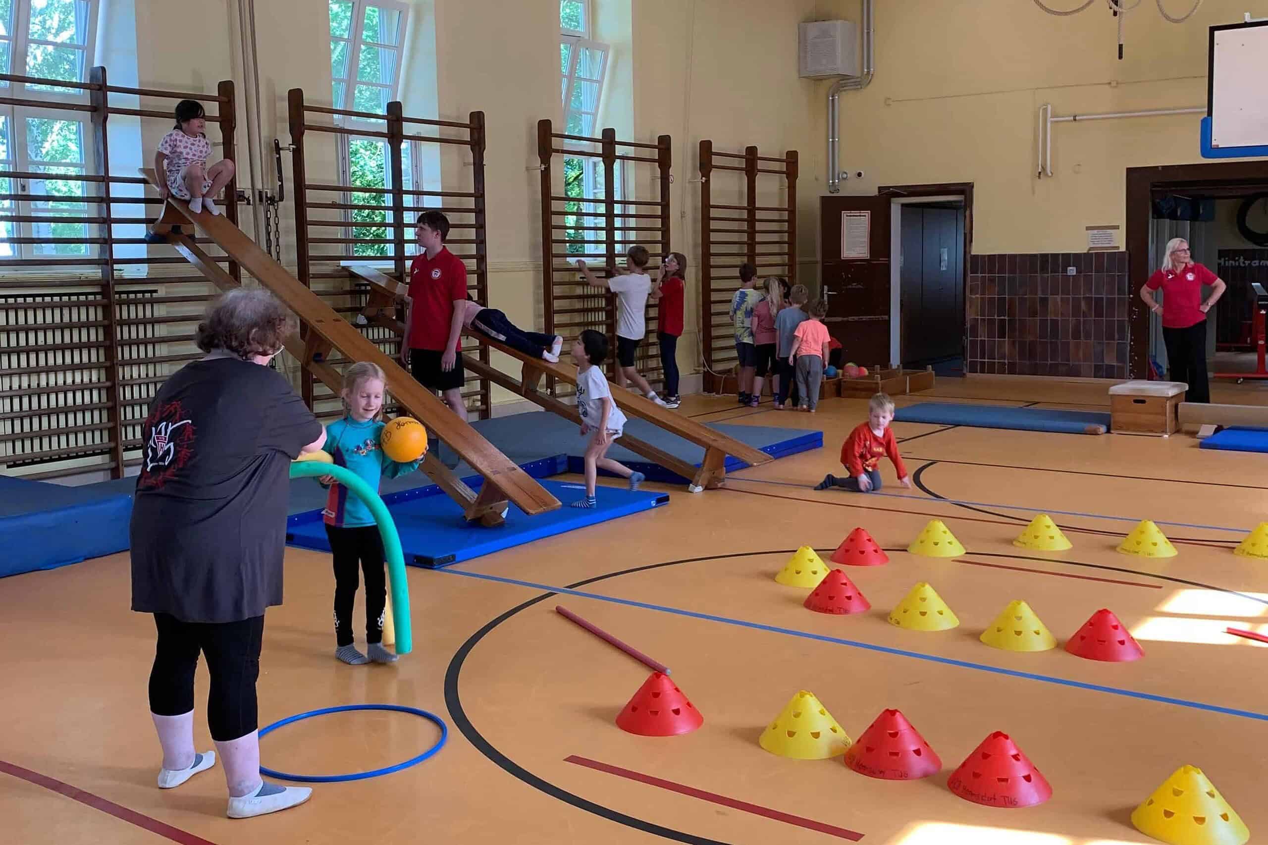 Kinder an Balancierbalken, Sprossenwänden und einem Hütchenparcours in einer Sporthalle