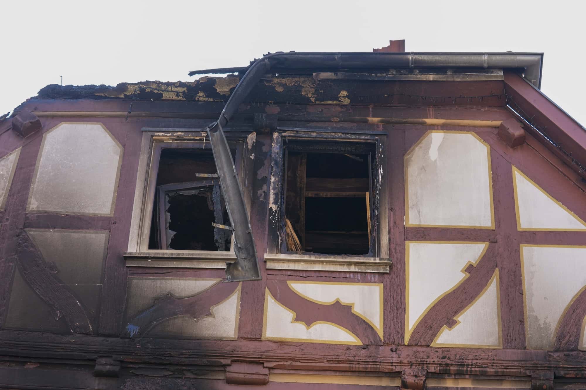 Detailaufnahme eines Fachwerkhauses mit rot-weiß bemalter Fassade: Das Dach ist abgebrannt, hinter zwei leeren Fensteröffnungen ist verkohltes Holz zu sehen.