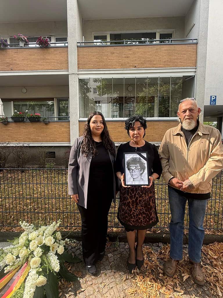 Drei Personen stehen vor einem Wohnhaus, die mittlere hält ein gerahmtes Porträt von Beate Fischer