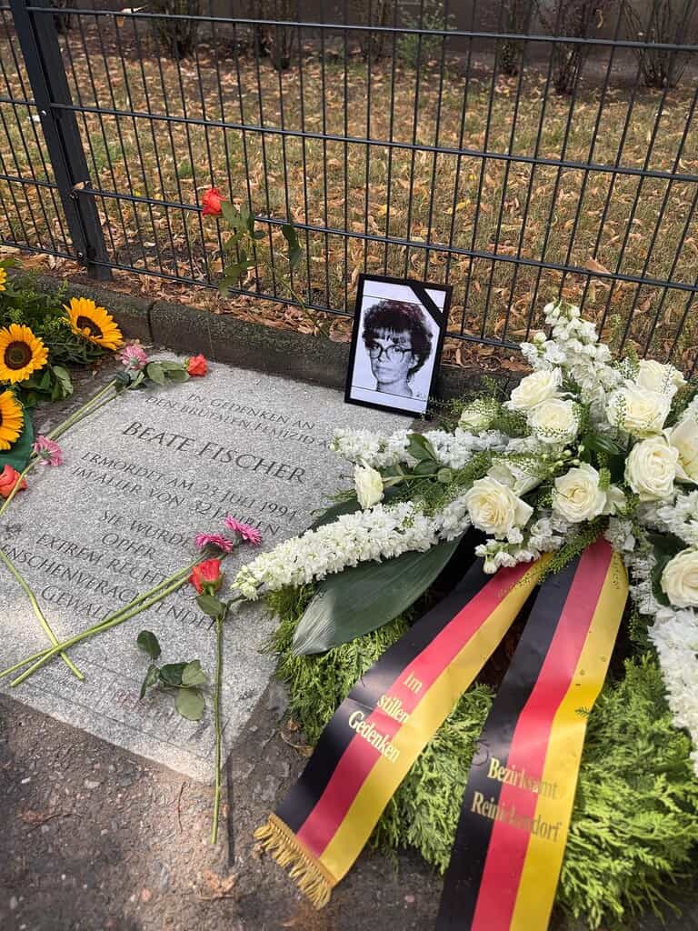 Gedenkplatte für Beate Fischer am Boden, daneben ein gerahmtes Porträt, Blumen und ein Trauerkranz
