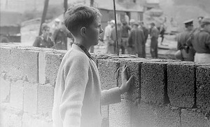Ein Junge steht vor einer halbhohen Mauer aus Hohlblocksteinen und legt die Hand darauf, dahinter arbeiten Männer an der Grenzbefestigung.
