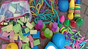 Bunte Spielgeräte wie Schwämme, Springseile, Wackelhalbkugeln und Wäscheklammern liegen auf einem Gehweg ausgebreitet.