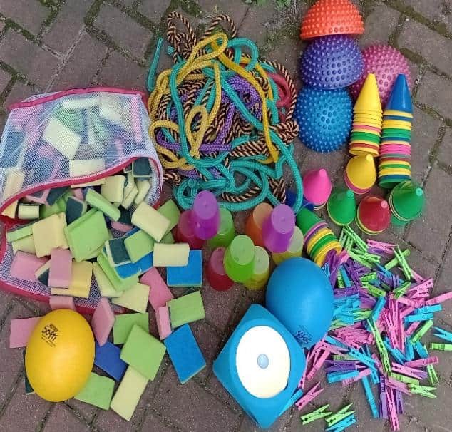 Bunte Spielgeräte wie Schwämme, Springseile, Wackelhalbkugeln und Wäscheklammern liegen auf einem Gehweg ausgebreitet.