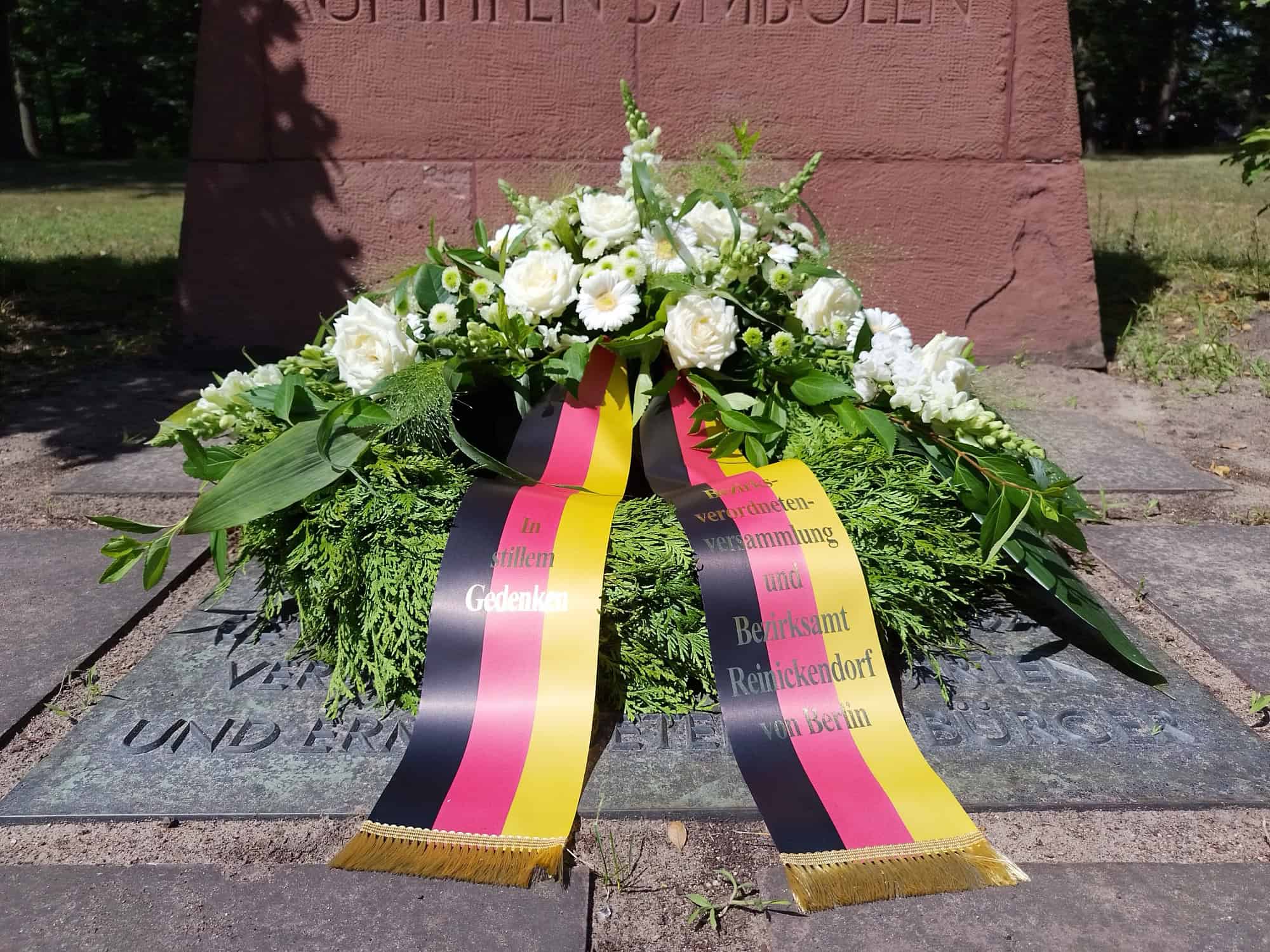 Kranz mit weißen Blumen und schwarz-rot-goldenen Schleifen der Bezirksverordnetenversammlung und des Bezirksamts Reinickendorf an einem Gedenkstein.