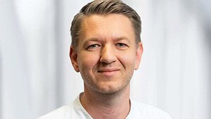 Porträt von Dr. med. Robert Steinicke im weißen Arztkittel über blauer Kasackjacke vor einem hellen, unscharfen Hintergrund.