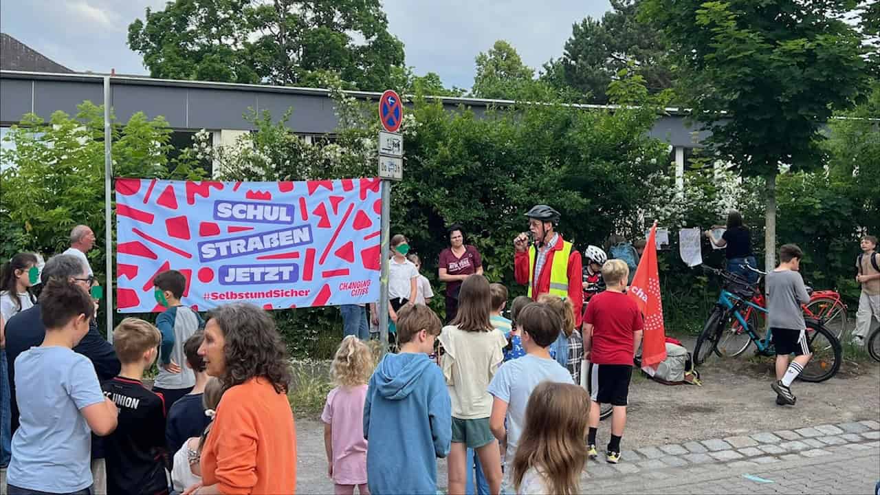 Ein Banner mit der Aufschrift Schulstraßen jetzt hängt an einem Gerüst, davor stehen Kinder und Erwachsene auf der Straße.