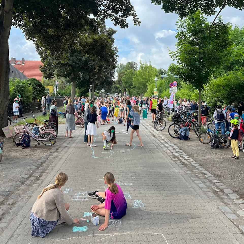 Zwei Mädchen sitzen auf dem Asphalt und malen mit Straßenkreide, im Hintergrund Fahrräder und eine Menschenmenge.