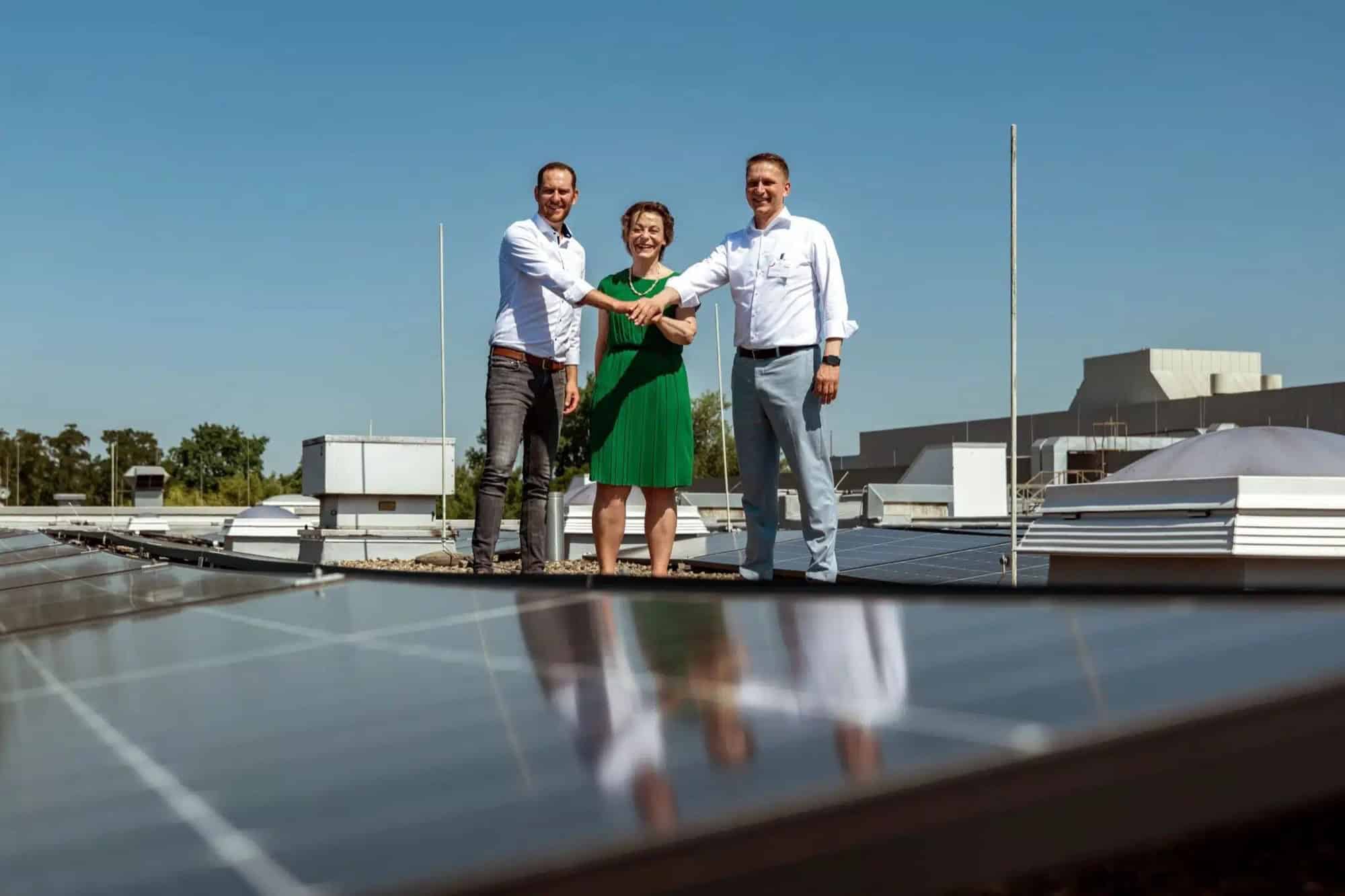 Drei Personen reichen sich auf einem Klinikdach zwischen Solarmodulen die Hände
