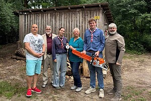 Sechs Personen stehen vor einer Holzhütte im Wald, eine Frau hält eine Plakette, ein junger Mann eine Motorsäge