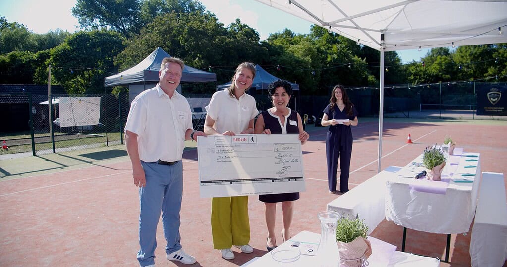 Drei Personen halten auf einem Tennisplatz einen großen symbolischen Scheck über 500 Euro in die Kamera.
