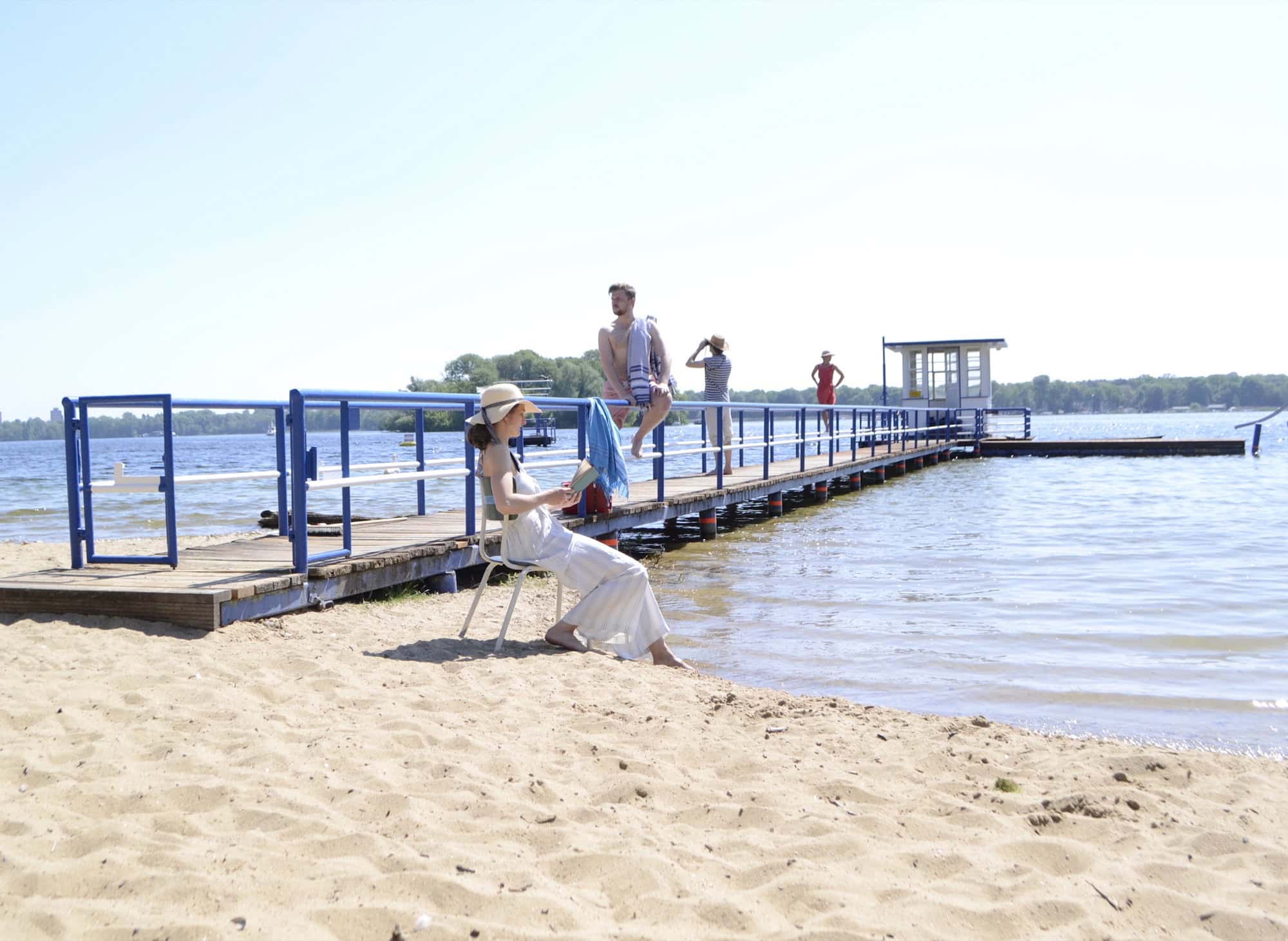 Eine Darstellerin im weißen Sommerkleid sitzt am Sandstrand neben einem Steg, im Hintergrund weitere Personen und der Tegeler See