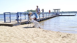 Eine Darstellerin im weißen Sommerkleid sitzt am Sandstrand neben einem Steg, im Hintergrund weitere Personen und der Tegeler See