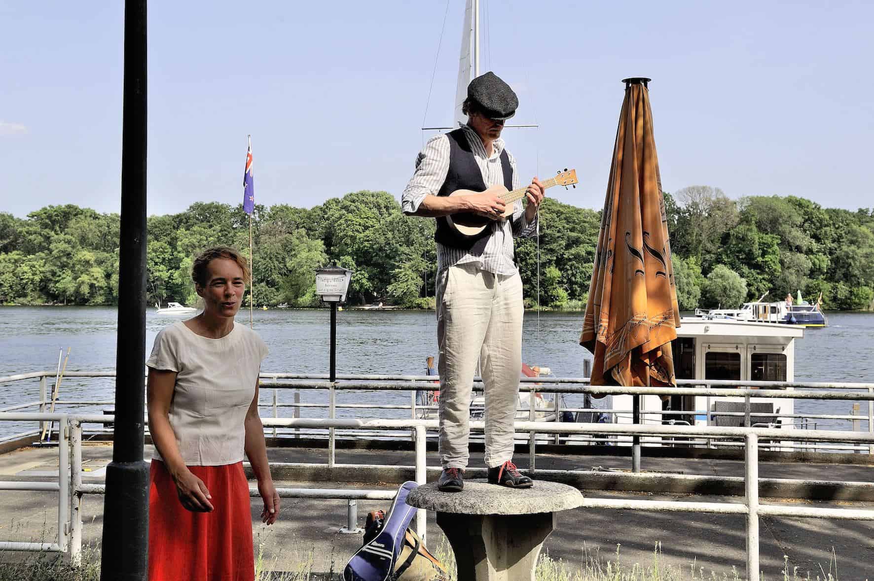 Ein Darsteller in heller Kleidung spielt auf einem Sockel stehend Ukulele, davor steht eine Darstellerin, im Hintergrund der Tegeler See