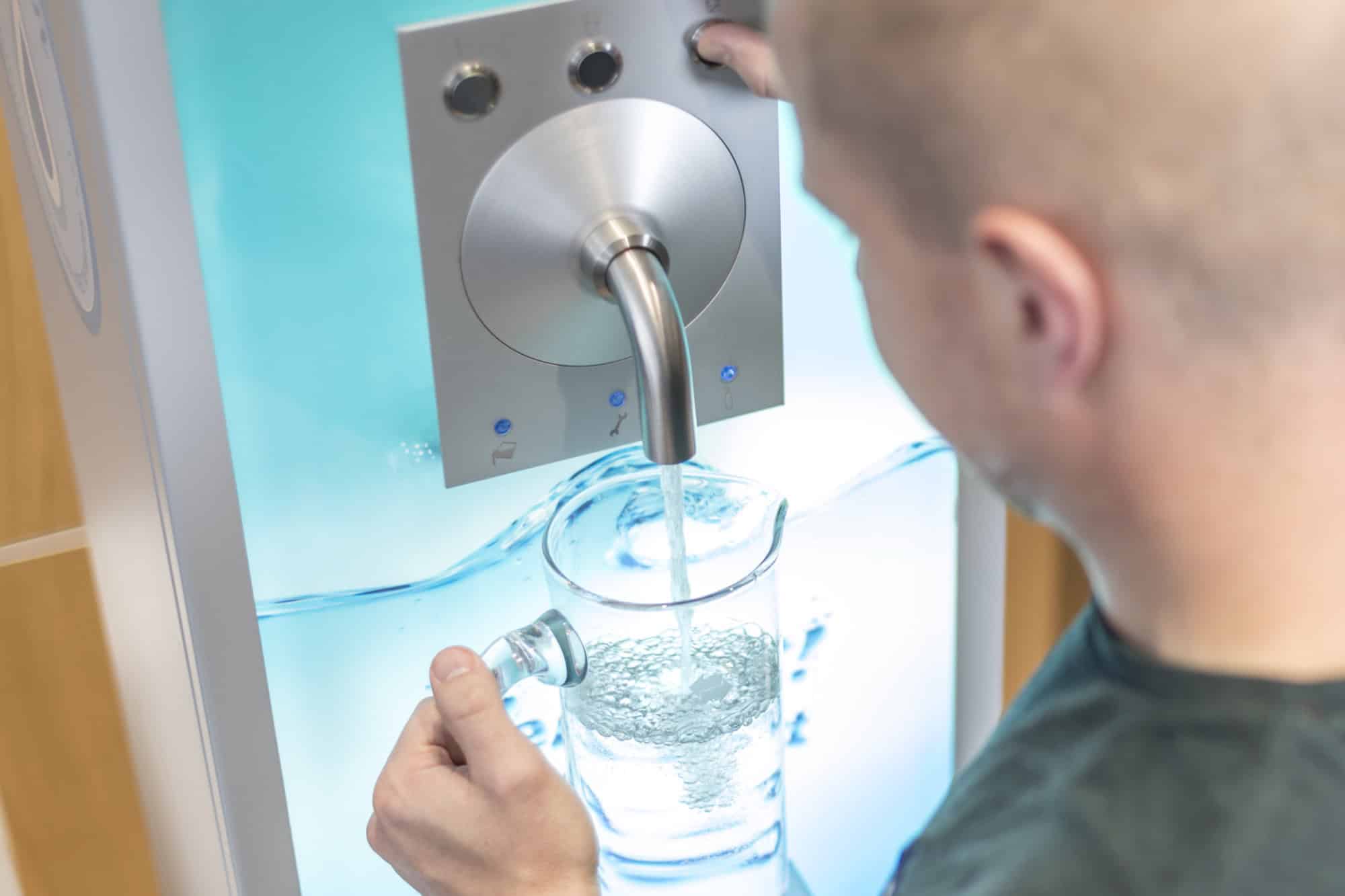 Ein Mann füllt an einem Wandwasserspender ein Glas mit sprudelndem Trinkwasser