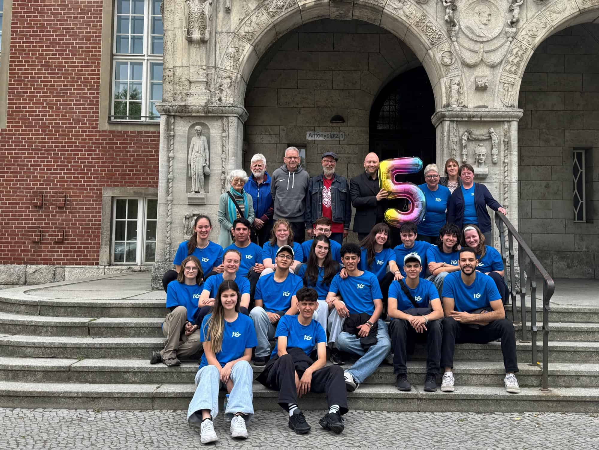 Gruppenbild mit 17 Jugendlichen in blauen T-Shirts und Erwachsenen auf einer Rathaustreppe, in der Mitte ein Luftballon in Form einer Fünf
