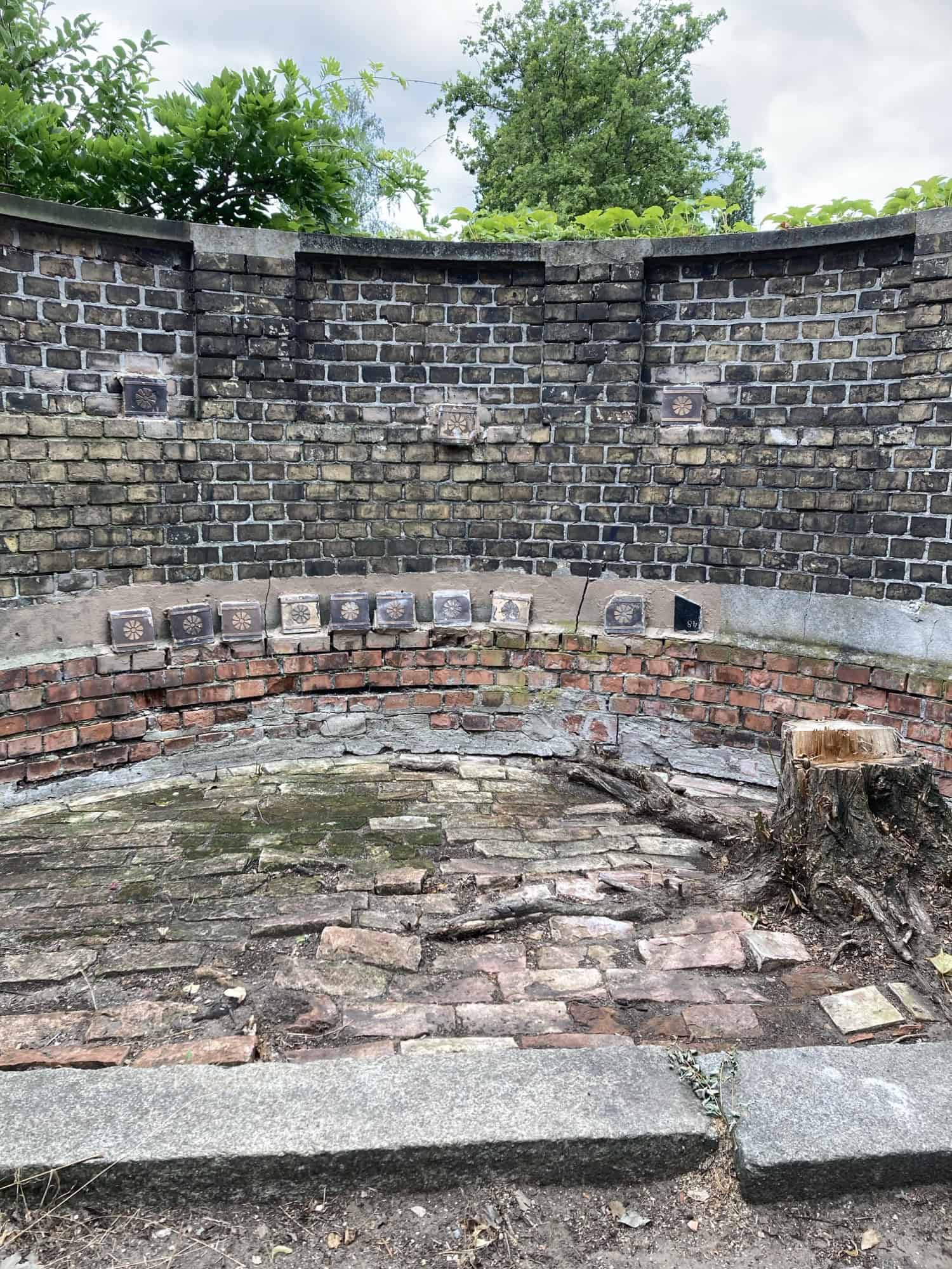 Freigelegtes Ziegelmauerwerk mit einer Reihe verzierter Kacheln auf dem Friedhofsgelände