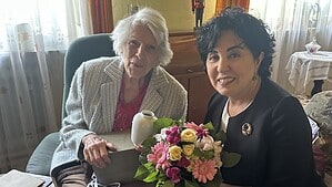 Eine ältere Frau mit weißem Haar sitzt in einem Sessel und hält eine Urkunde in den Händen, neben ihr sitzt eine Frau mit einem bunten Blumenstrauß auf dem Schoß.