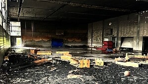 Blick in eine ausgebrannte Sporthalle: Der Boden ist mit schwarzen Trümmern und verkohlten Holzteilen bedeckt, die Decke ist rußgeschwärzt, an der Rückwand stehen rote Sitzbänke und eine blaue Matte.