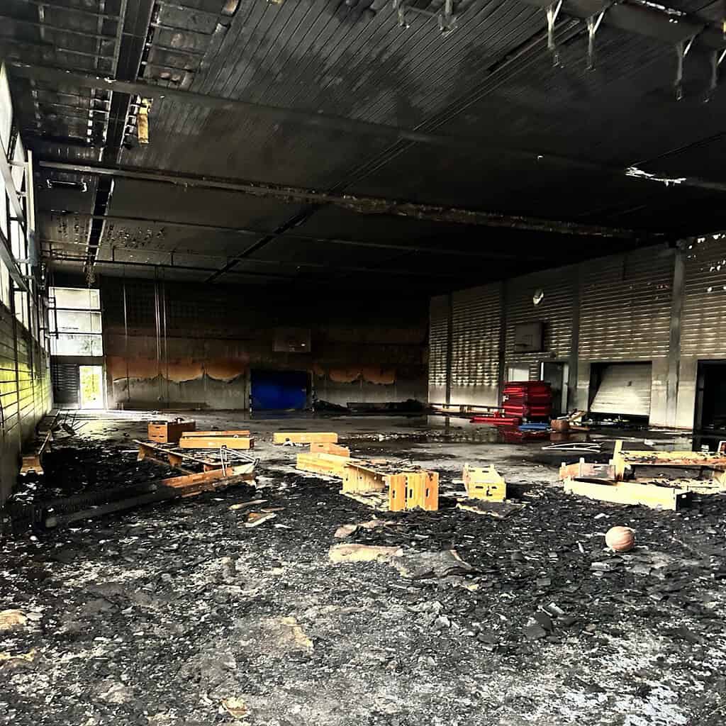 Blick in eine ausgebrannte Sporthalle: Der Boden ist mit schwarzen Trümmern und verkohlten Holzteilen bedeckt, die Decke ist rußgeschwärzt, an der Rückwand stehen rote Sitzbänke und eine blaue Matte.