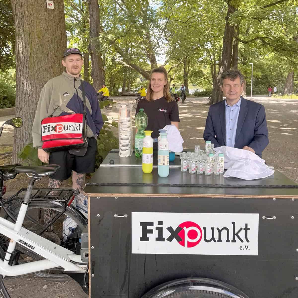 Zwei Mitarbeitende von Fixpunkt e. V. und Gesundheitsstadtrat Uwe Brockhausen stehen an einem Lastenrad mit Trinkflaschen, Bechern, Sonnencreme und Sonnenhüten unter Bäumen am Schäfersee.