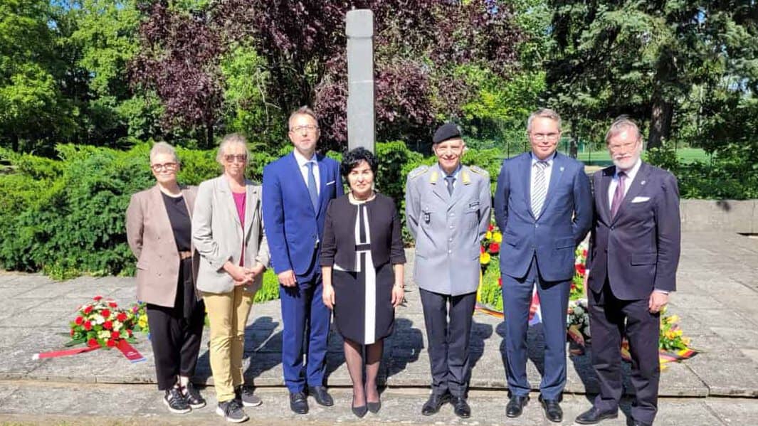 Sieben Personen in dunkler Kleidung und eine Person in Bundeswehruniform stehen vor einem hellen Steinmahnmal, davor liegen mehrere Kränze.