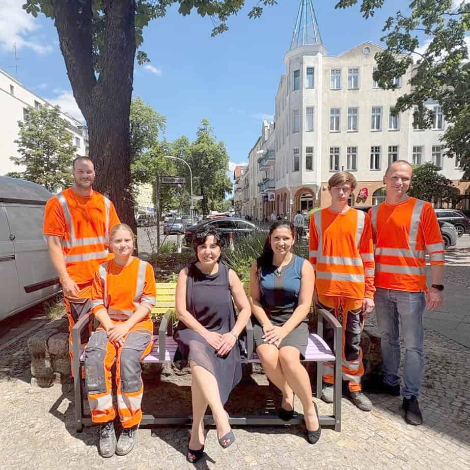 Eine bunt lackierte Bank auf einem Gehweg unter einem großen Baum, darauf sitzen zwei Frauen, umringt von vier Personen in orangefarbener Arbeitskleidung.