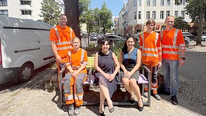 Eine bunt lackierte Bank auf einem Gehweg unter einem großen Baum, darauf sitzen zwei Frauen, umringt von vier Personen in orangefarbener Arbeitskleidung.
