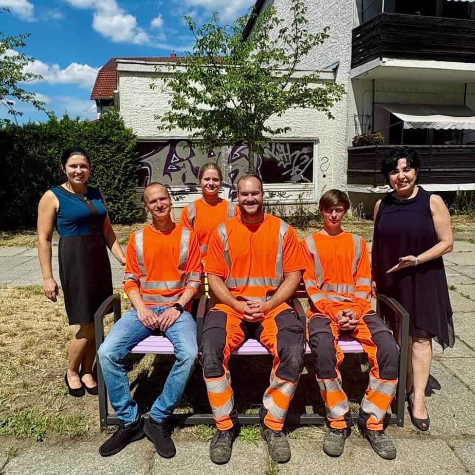 Eine bunt lackierte Bank auf einem Gehweg unter einem großen Baum, darauf sitzen zwei Frauen, umringt von vier Personen in orangefarbener Arbeitskleidung.