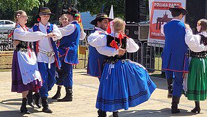 Tänzerinnen und Tänzer in bunten polnischen Trachten mit blauen Röcken und westenartigen Jacken tanzen paarweise vor einer Bühne im Freien.