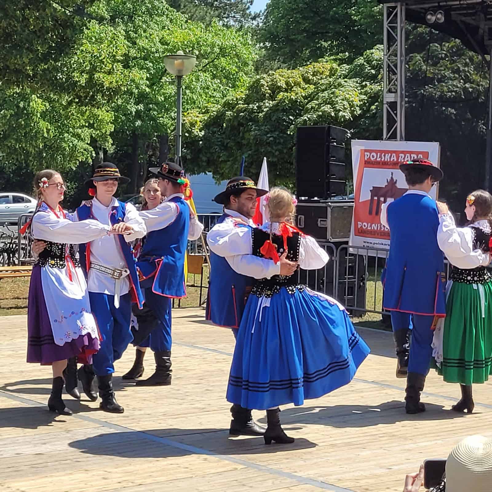 Tänzerinnen und Tänzer in bunten polnischen Trachten mit blauen Röcken und westenartigen Jacken tanzen paarweise vor einer Bühne im Freien.