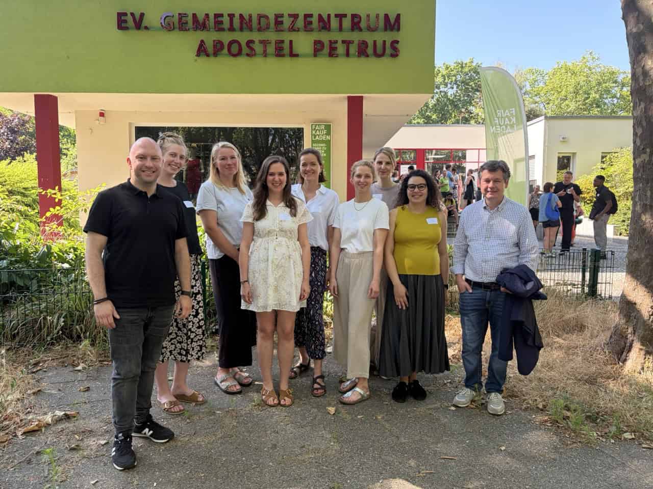 Zehn Personen stehen im Freien nebeneinander vor einem Gebäude mit der roten Aufschrift Ev. Gemeindezentrum Apostel Petrus.
