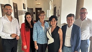 Sechs Personen stehen nebeneinander in einem hellen Flur mit Holzbalkendecke und Kronleuchtern und blicken in die Kamera.