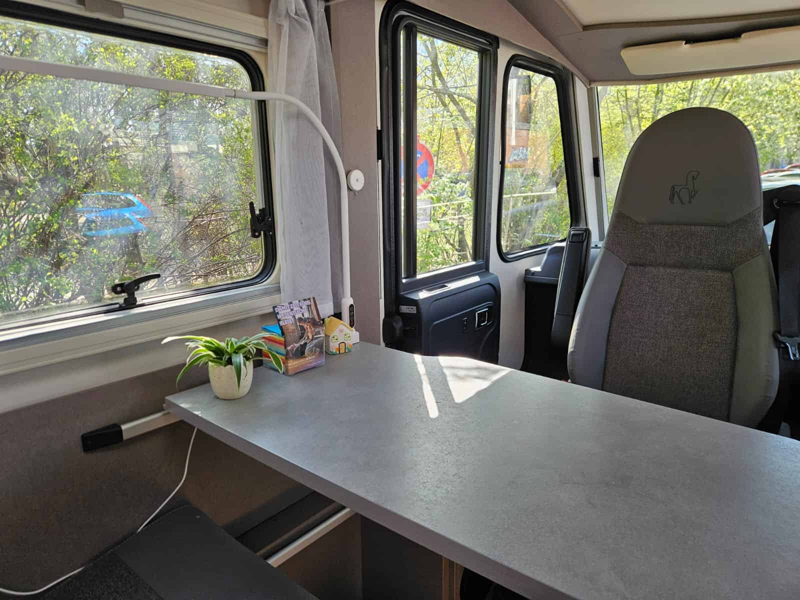 Innenraum des Beratungsbusses mit langem Tisch, Bürostuhl, Zimmerpflanze und Informationsmaterial, durch die Fenster sind Bäume zu sehen.