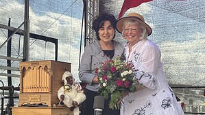 Zwei Frauen stehen auf einer Bühne neben einer hölzernen Drehorgel auf einem Wagen, die rechte trägt einen Strohhut und hält einen Blumenstrauß.