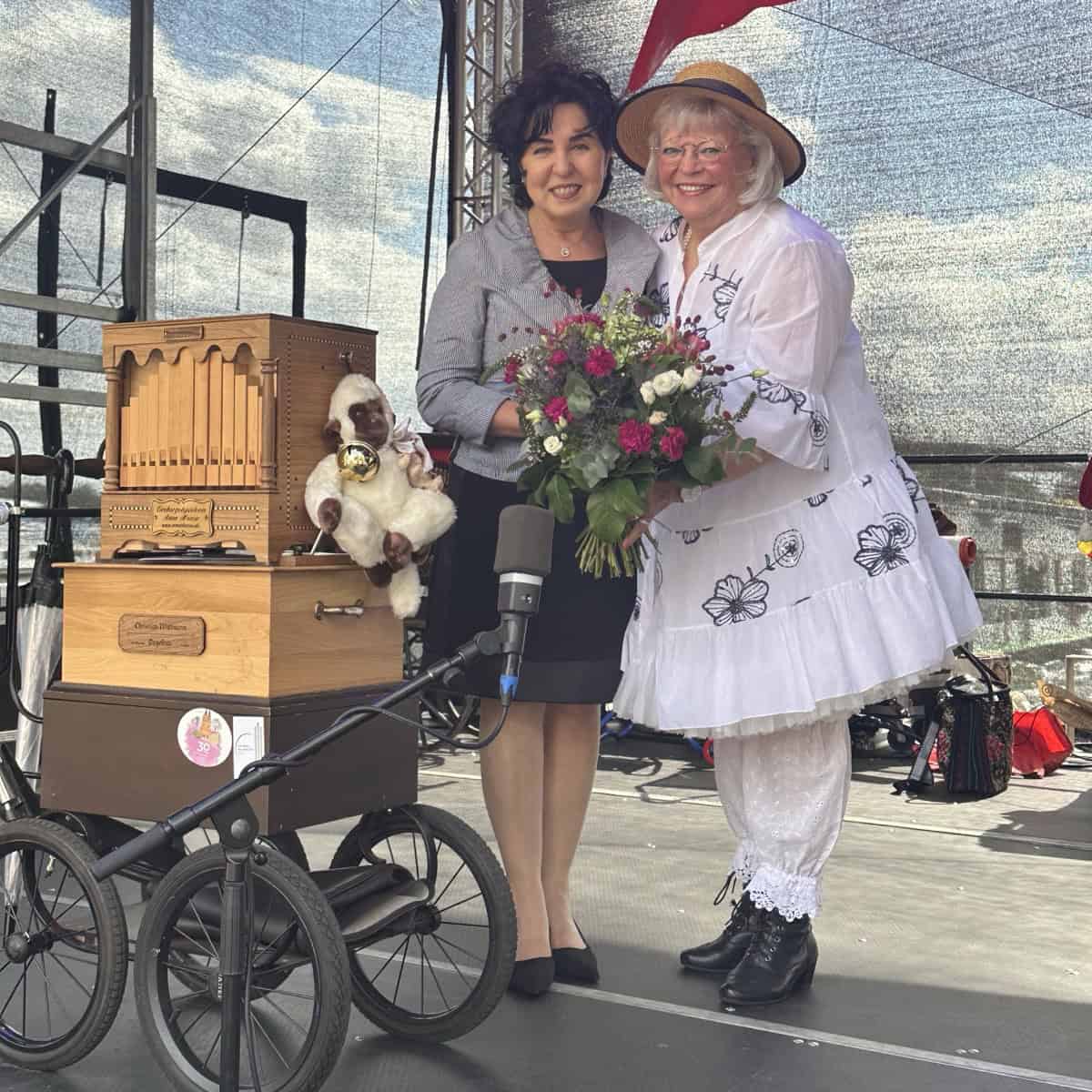 Zwei Frauen stehen auf einer Bühne neben einer hölzernen Drehorgel auf einem Wagen, die rechte trägt einen Strohhut und hält einen Blumenstrauß.