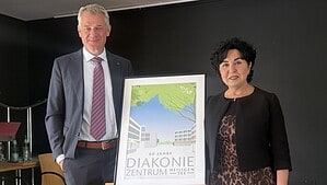 Eine Frau und ein Mann stehen in einem Saal links und rechts neben einer gerahmten Grafik mit der Aufschrift 60 Jahre Diakoniezentrum Heiligensee.