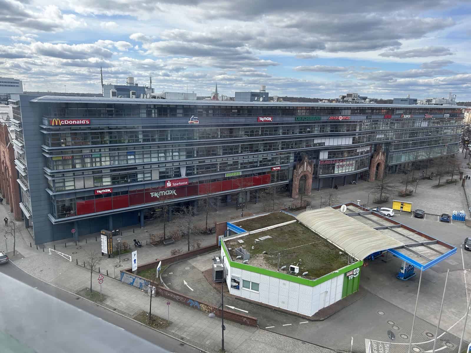 Blick von oben auf den langgestreckten, verglasten Gebäudekomplex der Hallen am Borsigturm mit Ladenschildern an der Fassade, davor eine Tankstelle und eine breite Straße.