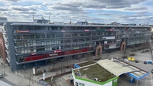 Blick von oben auf den langgestreckten, verglasten Gebäudekomplex der Hallen am Borsigturm mit Ladenschildern an der Fassade, davor eine Tankstelle und eine breite Straße.