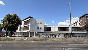 Langgestrecktes ehemaliges Kaufhausgebäude mit heller Plattenfassade und leeren Schaufenstern an einer breiten Straße, davor eine Bushaltestelle und Bäume.