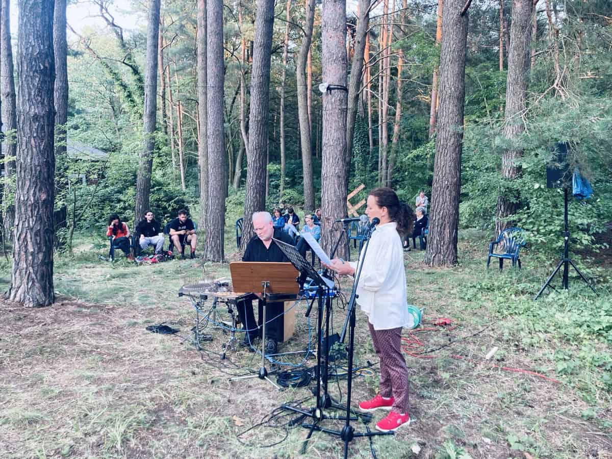 Eine Sprecherin am Mikrofon und ein Musiker an einem Instrumententisch treten im Wald vor sitzendem Publikum auf